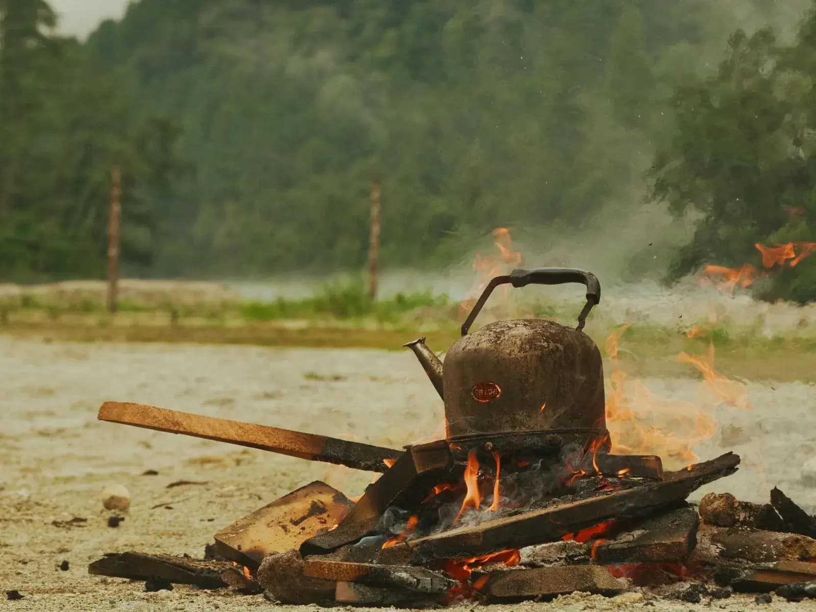 A soot-blackened kettle heating on an open wood fire with a misty forested mountain behind
