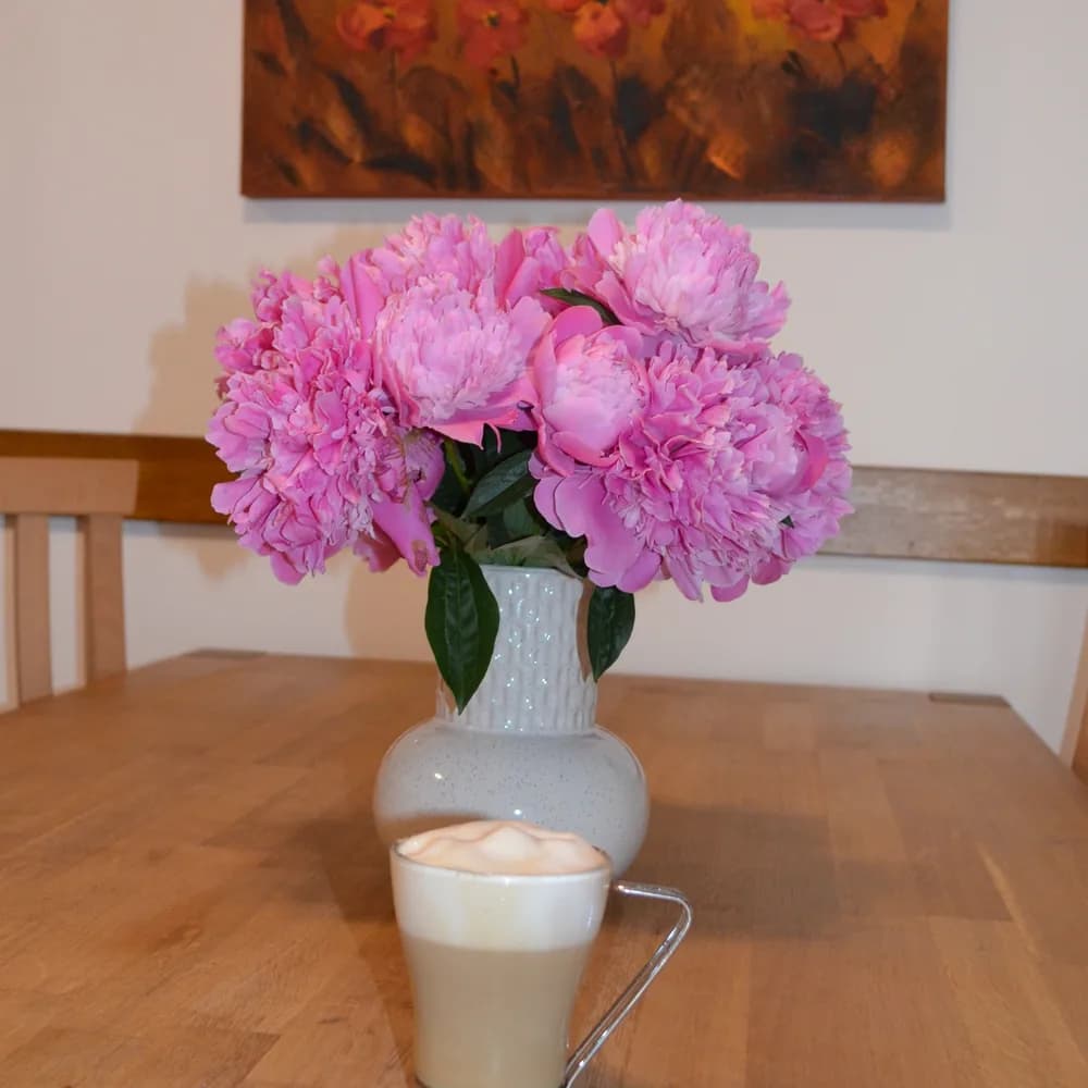 A jug of pink peonies on the farmhouse dining table, a poppy painting on the wall behind