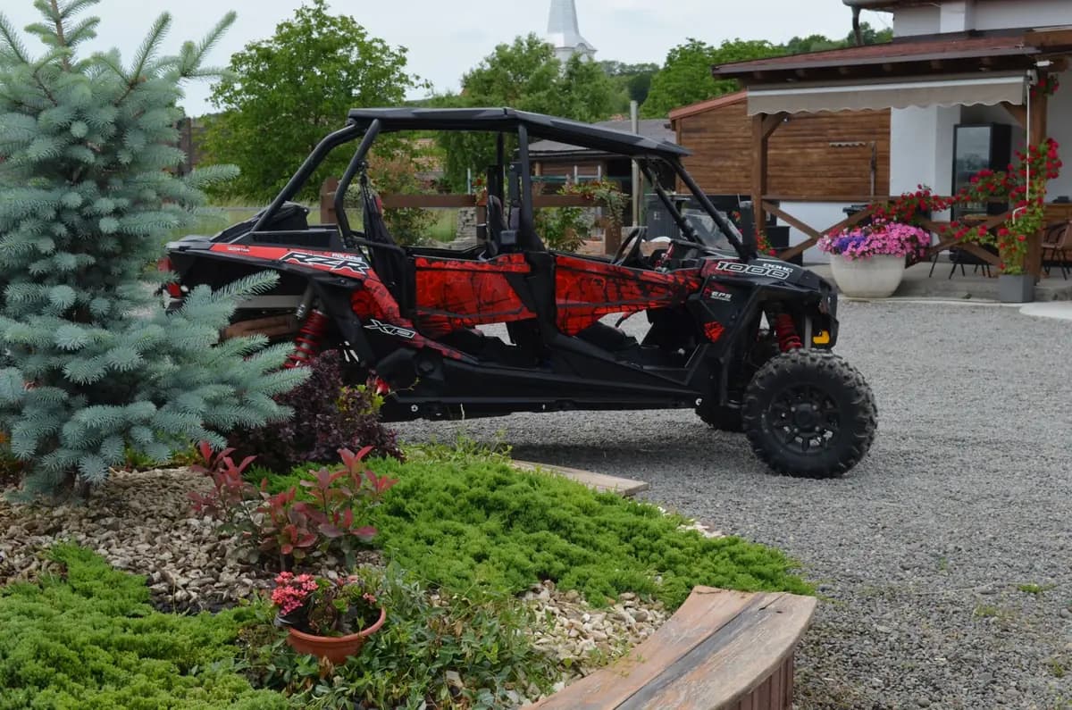A red and black four-seat Polaris RZR XP 1000 side-by-side parked on the gravel drive beside the farmhouse