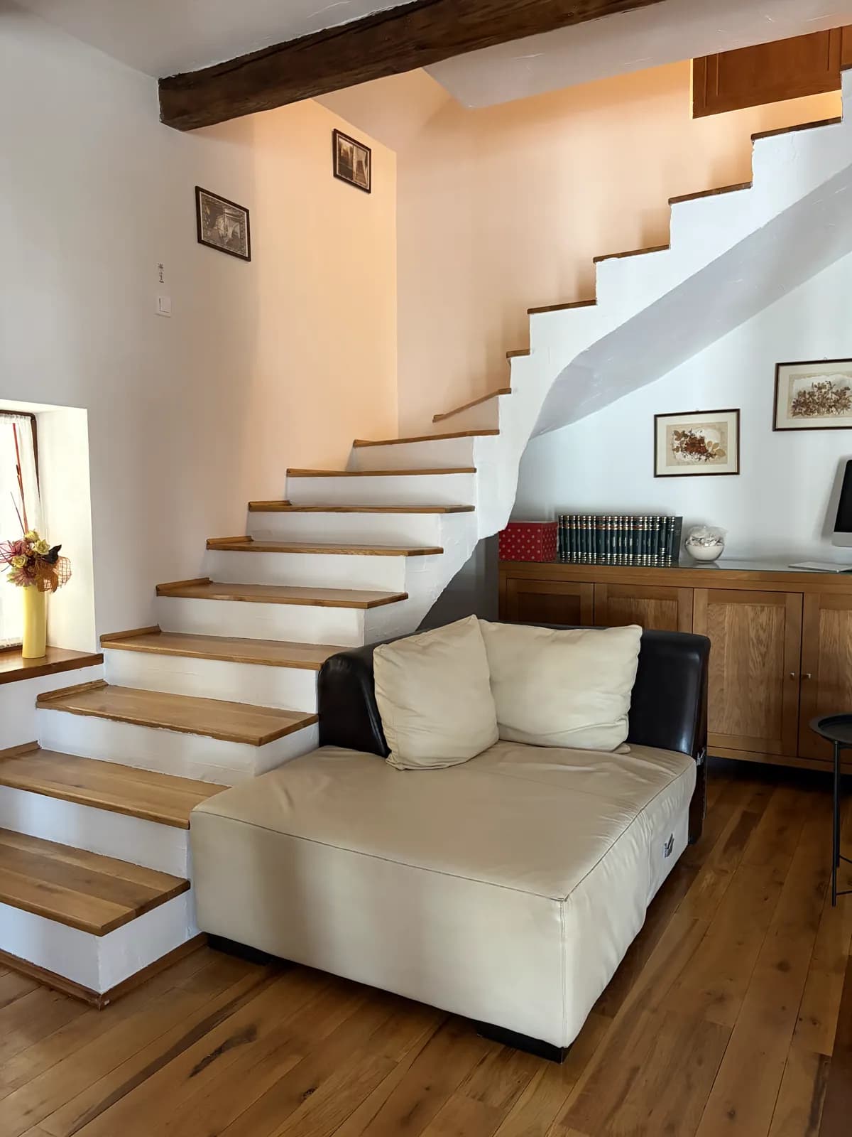 A white staircase with oak treads curving up past a cream leather sofa and a low wooden cabinet of books