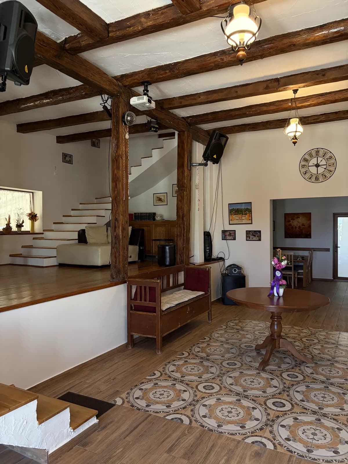 A wood-beamed hall with a raised lounge and staircase to one side, a round table on a patterned tile floor and a wall clock