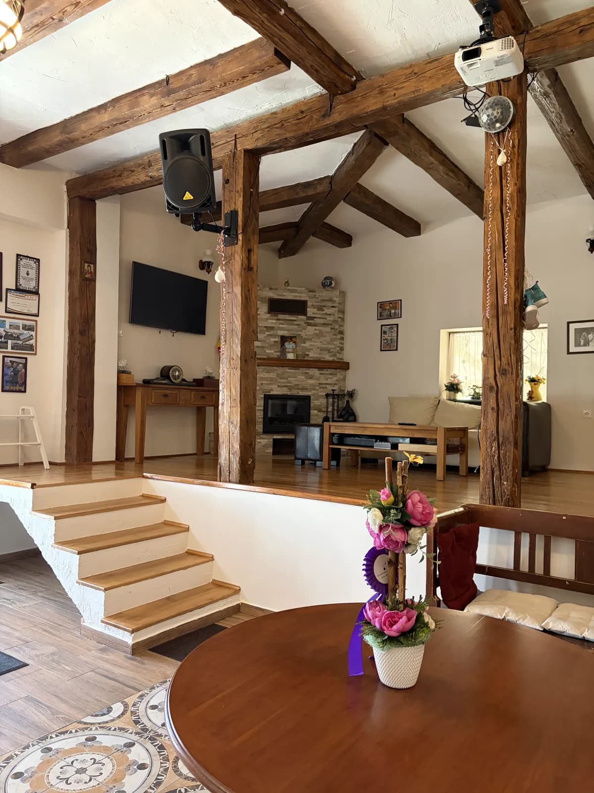 A round wooden table with flowers in the foreground, steps rising to a beamed lounge with a stone fireplace and TV