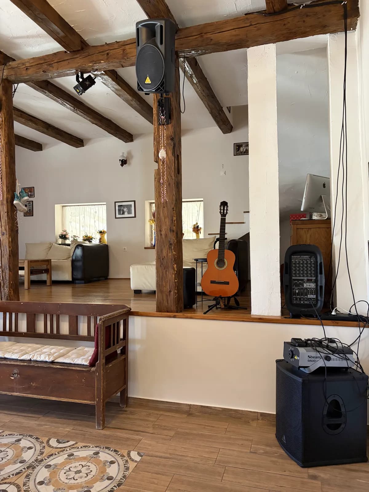 An acoustic guitar on a stand and party speakers on a raised timber-beamed floor, a painted wooden bench below
