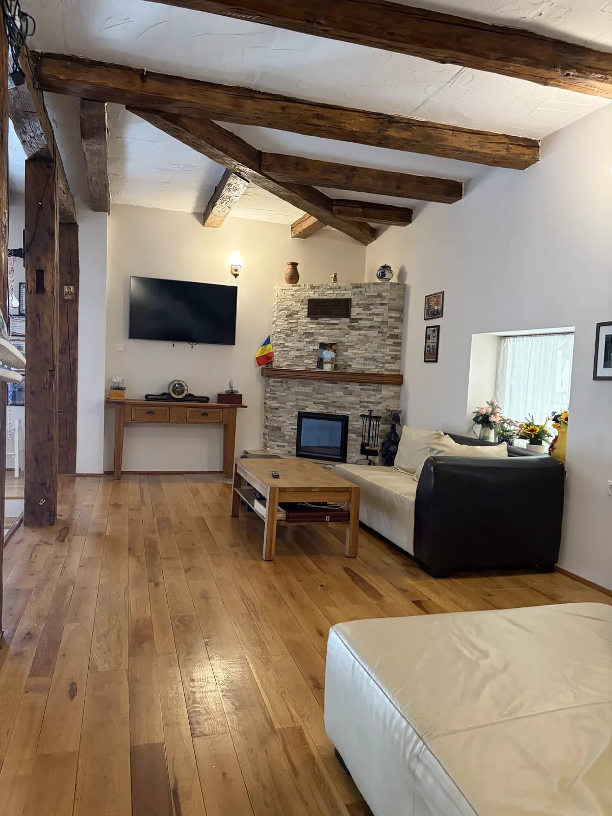 A living room with exposed timber beams, a stone fireplace, a wall-mounted TV and leather sofas