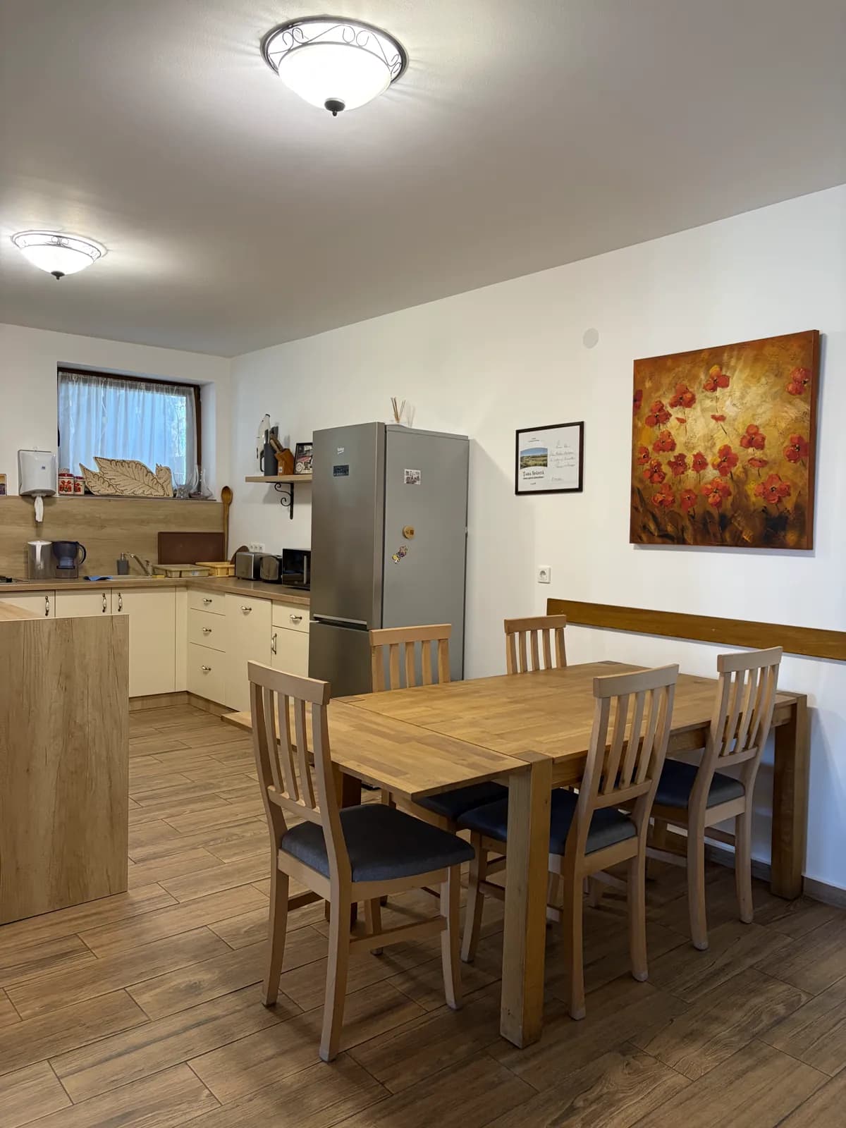 An open kitchen and dining area with a fridge, a long wooden table and chairs, a poppy painting on the wall