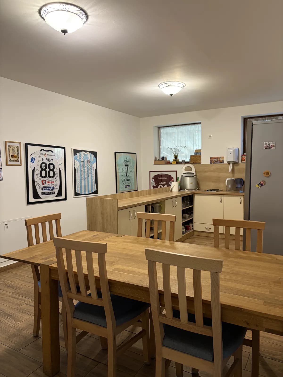 A long wooden dining table and chairs in front of the kitchen counter, framed signed football shirts on the wall