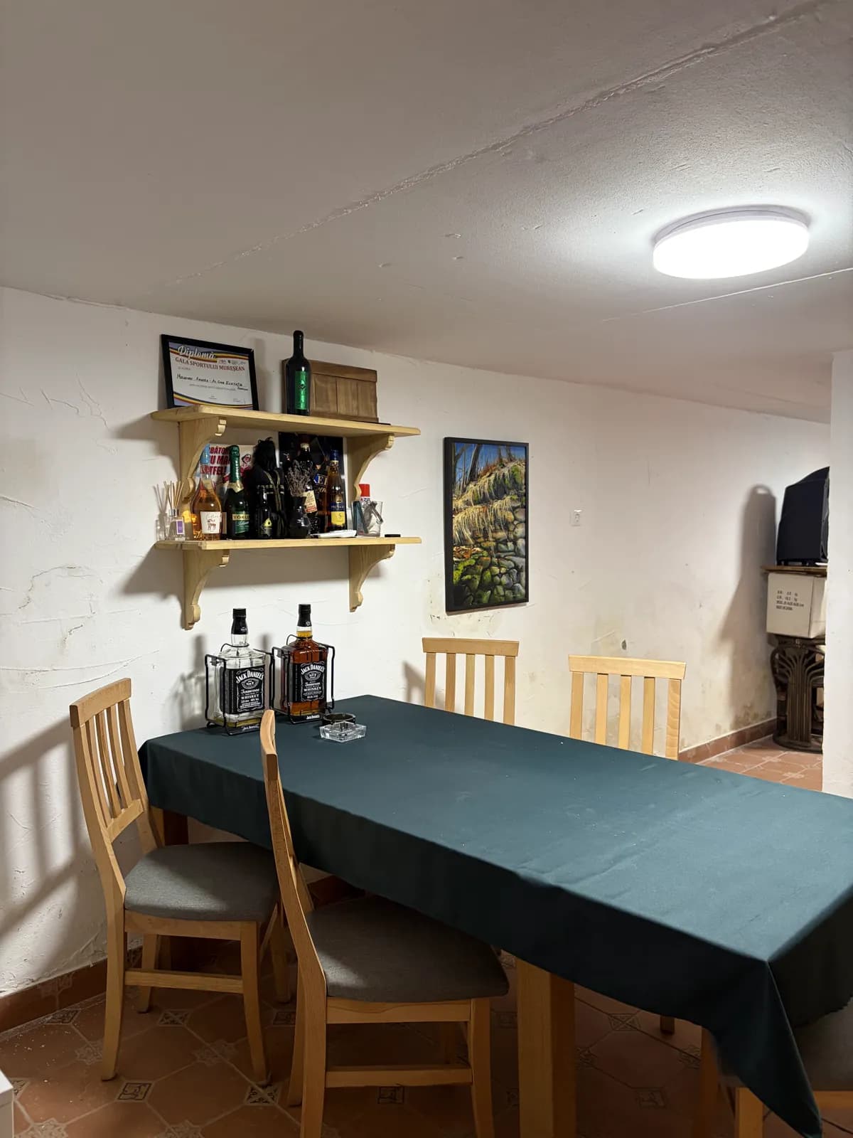 A dining table with a green cloth in a whitewashed cellar room, a shelf of bottles on the wall