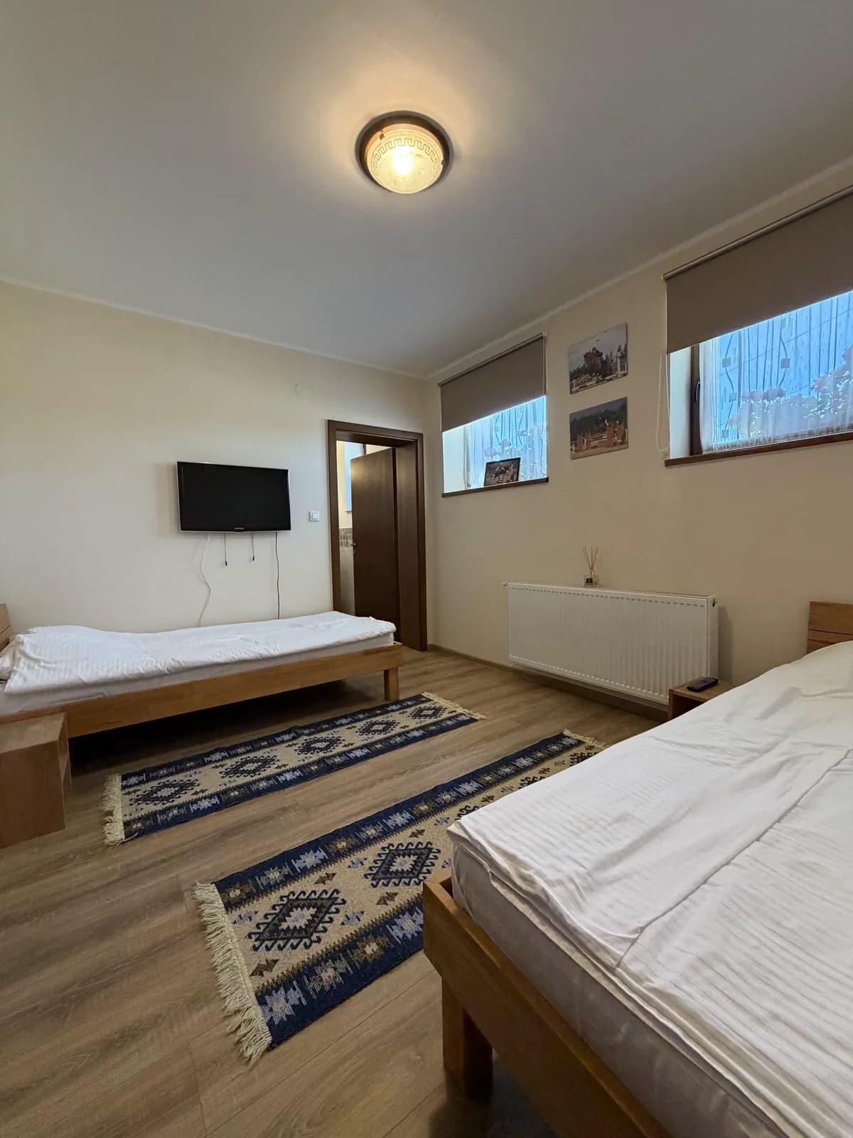 A ground-floor room with two single beds, a wall-mounted TV, patterned kilim runners on the floor and framed photographs between the windows