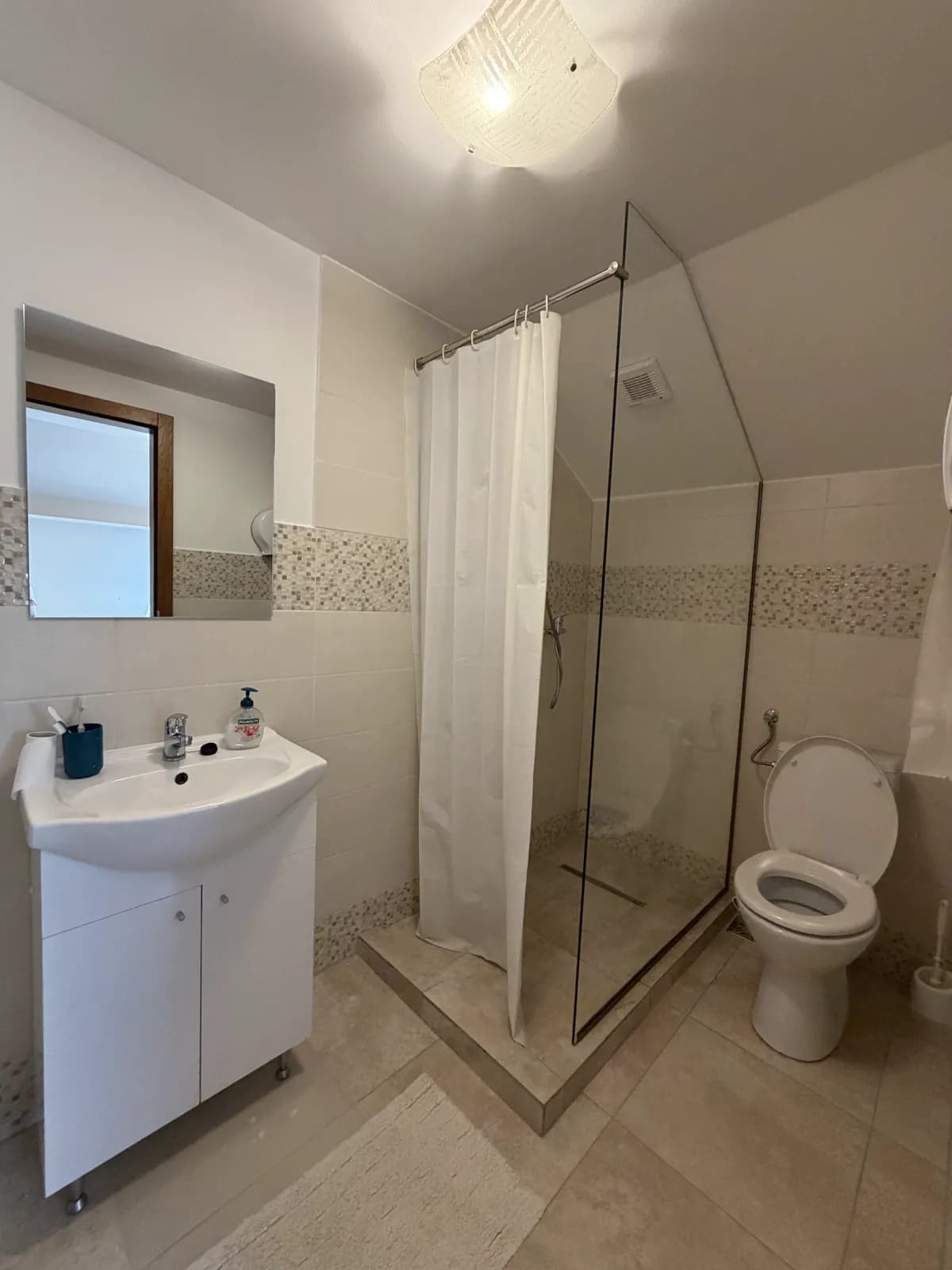 A cream-tiled attic bathroom under a sloped ceiling, with a glass shower, sink and toilet