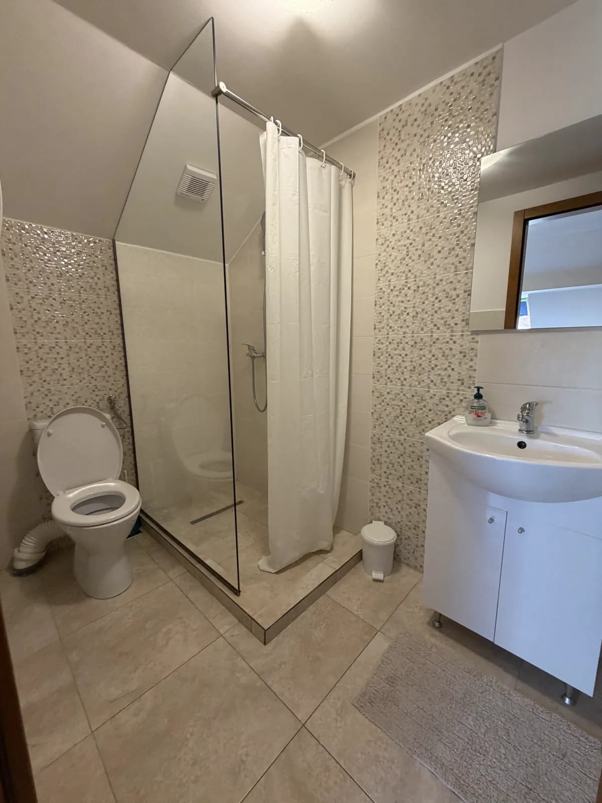 A cream mosaic-tiled attic bathroom with a toilet, a glass shower stall and a sink