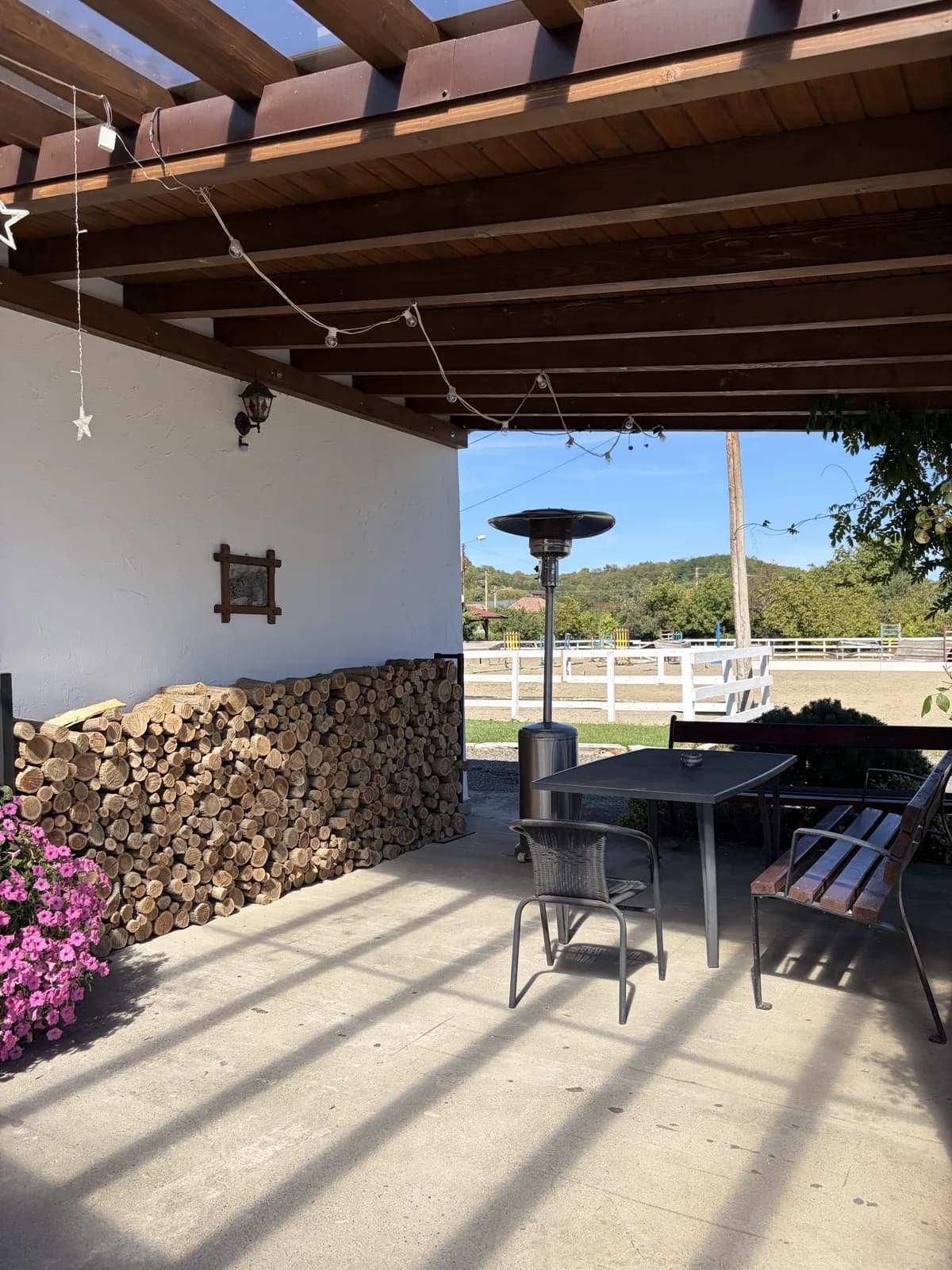 A covered terrace with a stacked woodpile, a patio heater, a table and bench, and the white-fenced riding paddock beyond