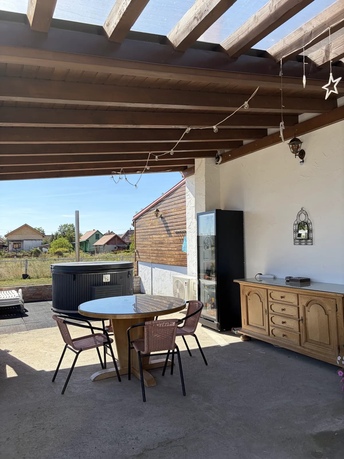 A round dining table under the covered terrace, string lights overhead, a drinks fridge and the hot tub at the edge
