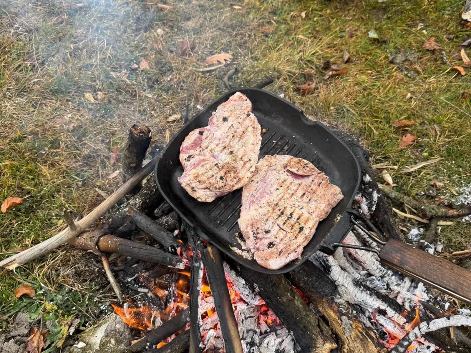 Two steaks grilling on a cast-iron pan over a campfire