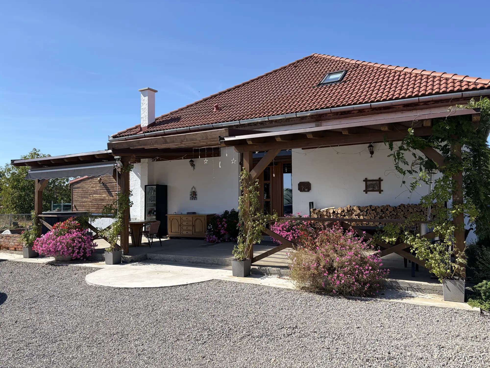 The whitewashed farmhouse with a terracotta roof and skylight, its timber pergola and pink flowering shrubs across the gravel yard