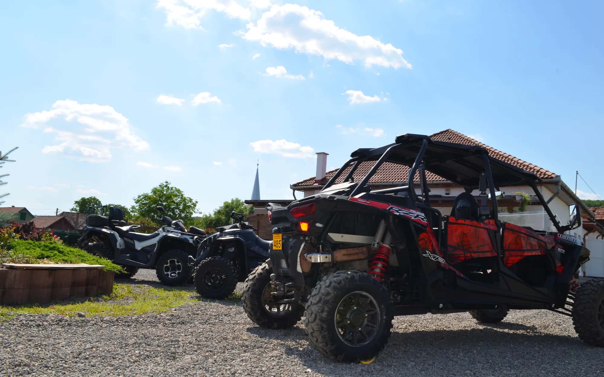 Two red-and-black RZR side-by-sides parked nose to tail in the gravel yard beside a wooden hay barn