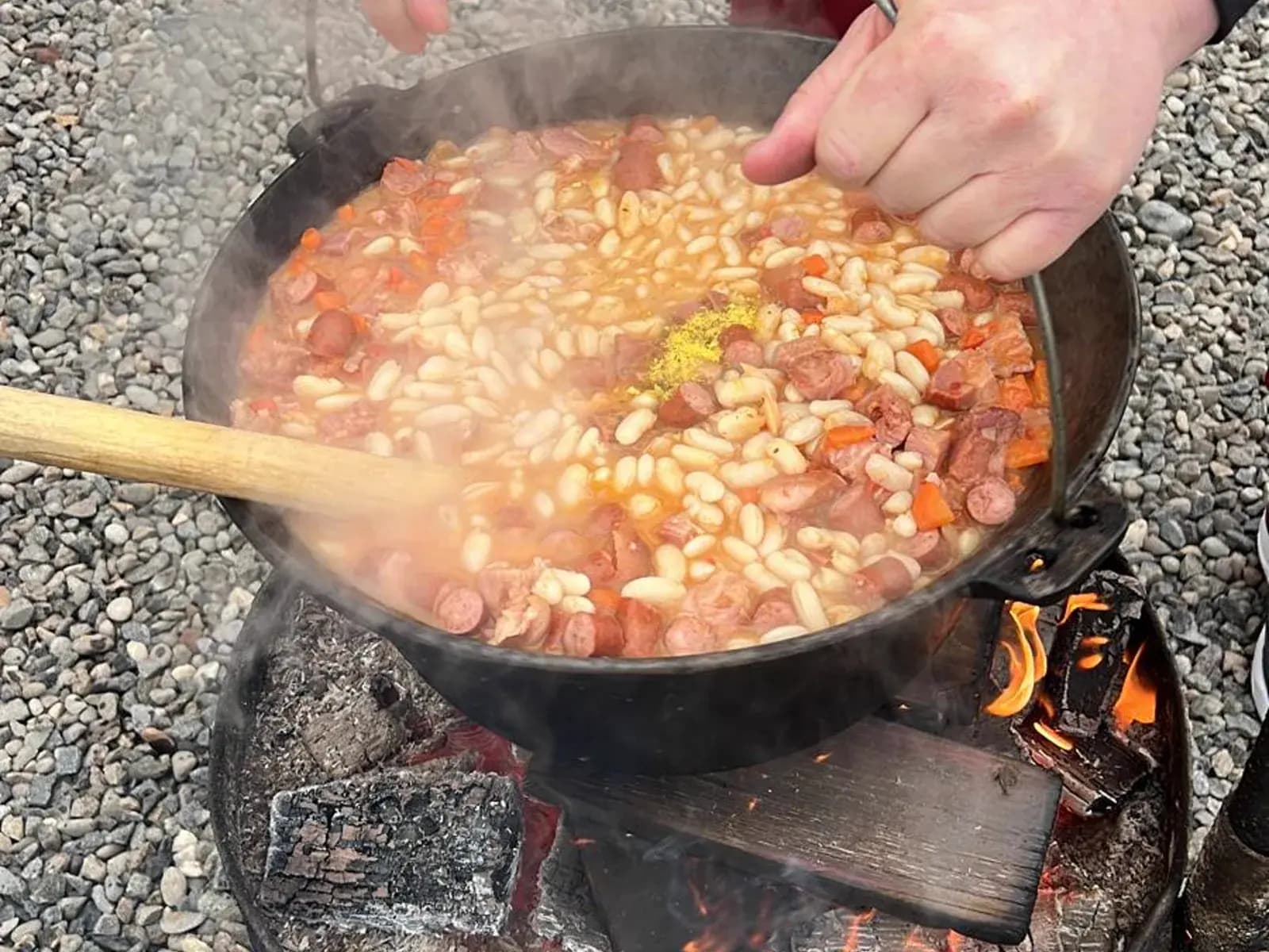 A traditional bean stew steaming in a cast-iron cauldron over an open fire