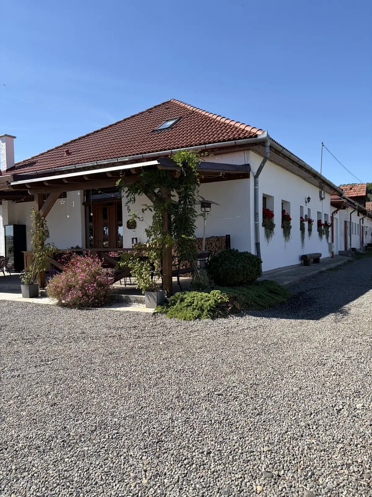 The farmhouse's corner and long whitewashed wing under a terracotta roof, red geraniums in the windows and a gravel yard in front